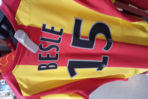 Ein rotes und gelbes Fußballtrikot mit Text und Nummern darauf, das an einer Stange in einem Geschäft hängt, zusammen mit anderen Gegenständen und einem sichtbaren Boden im Hintergrund.