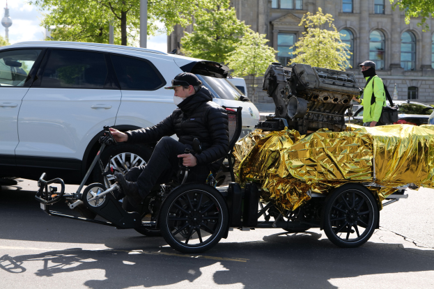 Ein Mann im Rollstuhl mit einem großen Motor auf dem Rücken, umgeben von Fahrzeugen auf einer Straße mit Bäumen, Gebäuden, Polen und einem klaren blauen Himmel im Hintergrund, trägt eine schwarze Jacke, eine Mütze und hält ein Objekt.