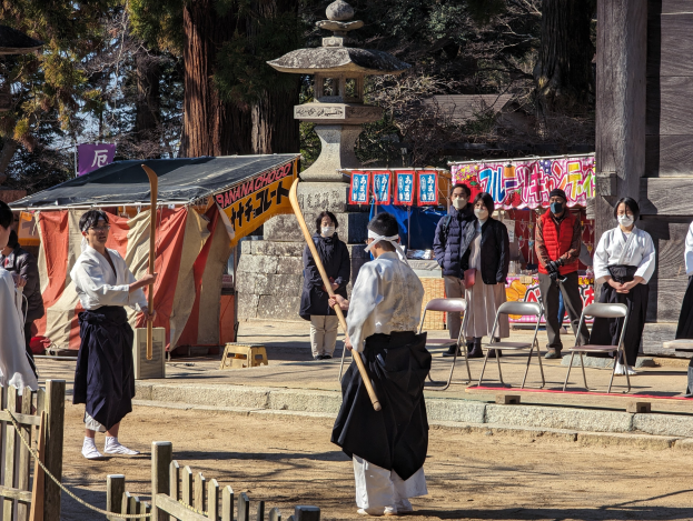 Eine Gruppe von Menschen in traditioneller Kleidung steht eng zusammen vor einem Gebäude in Kyoto, einige tragen Masken und halten Holzstöcke, mit Stühlen, Bannern, einem Zelt und Bäumen unter einem klaren blauen Himmel.