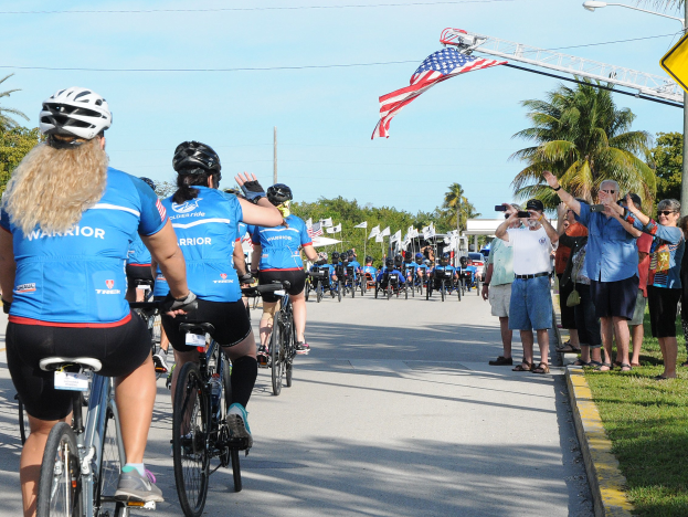 Radfahrer mit Helmen fahren eine von Bäumen gesäumte Straße während des 2017 Florida Triathlons, während Zuschauer auf der rechten Seite das Ereignis mit Kameras und Handys aufnehmen.