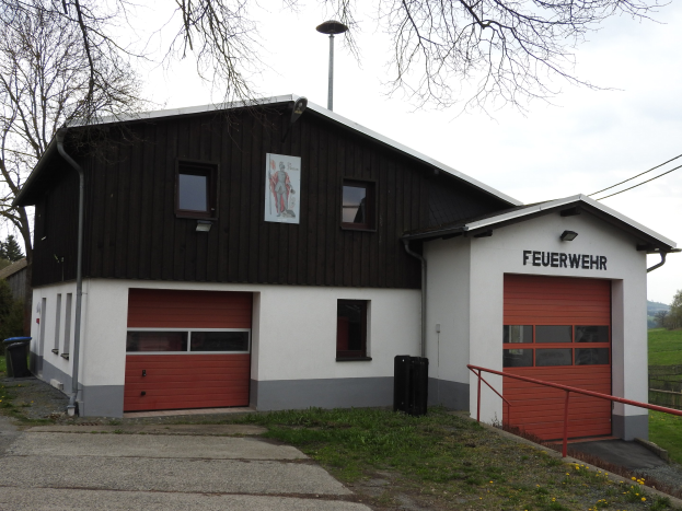 Feuerwache mit rotem und weißem Gebäude, Straße, Gras, gelben Blumen, schwarzen Objekten, Geländer, Pfahl, Drähte, Bäume und Himmel im Hintergrund.