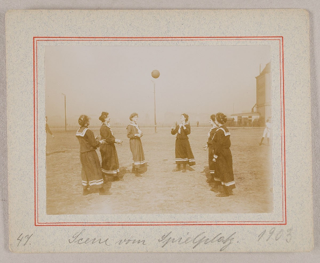 Eine Gruppe von Frauen, die Fußball auf dem Boden spielen, mit einem Ball in der Mitte, Pfosten und Gebäuden im Hintergrund und Text am unteren Rand des Bildes.