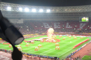 Ein Stadion mit Flutlicht, Eisengittern, Zuschauern, stehenden Personen, Werbetafeln, Anzeigebildschirmen, Sicherheitskameras, Fahnen und Dekorationen.