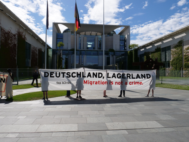 Gruppe von Menschen mit einem Banner "Deutschland, Lagerland Migration ist kein Verbrechen" vor einem Gebäude mit Bäumen, einem Metallzaun und Fahnen auf einer grasbewachsenen Fläche unter einem bewölkten Himmel.