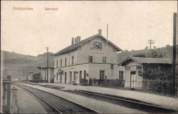 Schwarzes und weißes Foto einer Bahnstation in Dielkirchen, Deutschland, mit einem Zug auf den Schienen, umgeben von Häusern, Strommasten, Bäumen und Menschen, vor einem Hügel- und klaren Himmel.