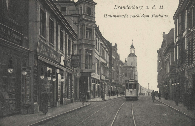 Schwarze und weiße Fotografie einer Stadtstraße in Brandenburg, Deutschland, mit einer Straßenbahn, Fußgängern, Gebäuden, Schildern, Straßenlaternen und einem bewölkten Himmel.