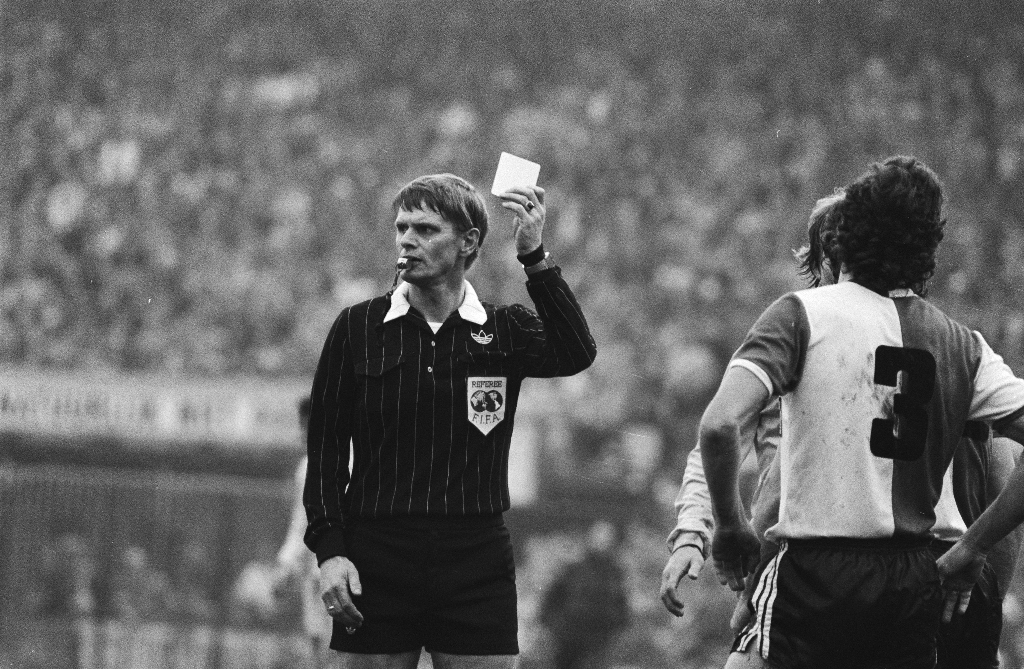 Schwarzes und weißes Bild eines Schiedsrichters mit roter Karte vor einer Menge in Fußballuniformen mit einem Zaun im Hintergrund.