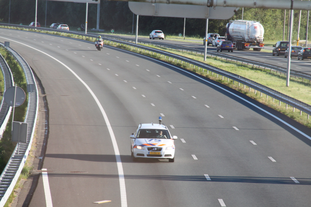 Ein Auto auf einer Autobahn mit einem stationären Polizeiwagen am Rand, andere Fahrzeuge und Bäume, Geländer, Pfosten und Planken im Hintergrund.