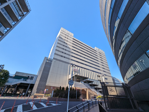 Hohes Gebäude mit zahlreichen Fenstern, identifiziert als Tokyo Metropolitan Government Building, in einer städtischen Umgebung mit Straßeninfrastruktur und wolkenlosem Himmel.