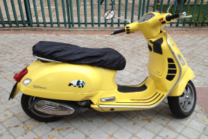 Ein gelbes Vespa GTS 300 Super ABS-Scooter steht auf der Straße geparkt, mit einem schwarzen Tuch bedeckt, mit einem Geländer, Gras und einigen Menschen im Hintergrund.