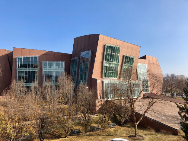 Großes Gebäude mit zahlreichen Fenstern, umgeben von Bäumen, Bänken, Gras und einem Laternenmast, mit anderen Gebäuden und einem klaren blauen Himmel im Hintergrund, identifiziert als der neue Campus der University of Illinois at Chicago.