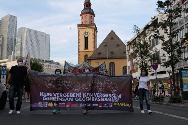 Eine Gruppe von maskierten Menschen, die eine Straße entlanggehen und ein Transparent mit Text und Bildern halten, mit einem geparkten Auto auf der linken Seite, Gebäuden, Bäumen, Schildern, Pfosten, einem Uhrenturm und einem klaren blauen Himmel im Hintergrund.