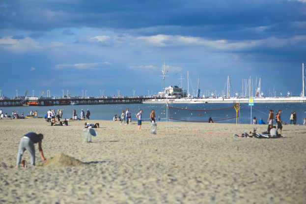 Gruppe von Menschen, die Volleyball auf einem sandigen Strand mit einem Netz spielen, mit Booten und einer Brücke im Hintergrund unter einem bewölkten Himmel.