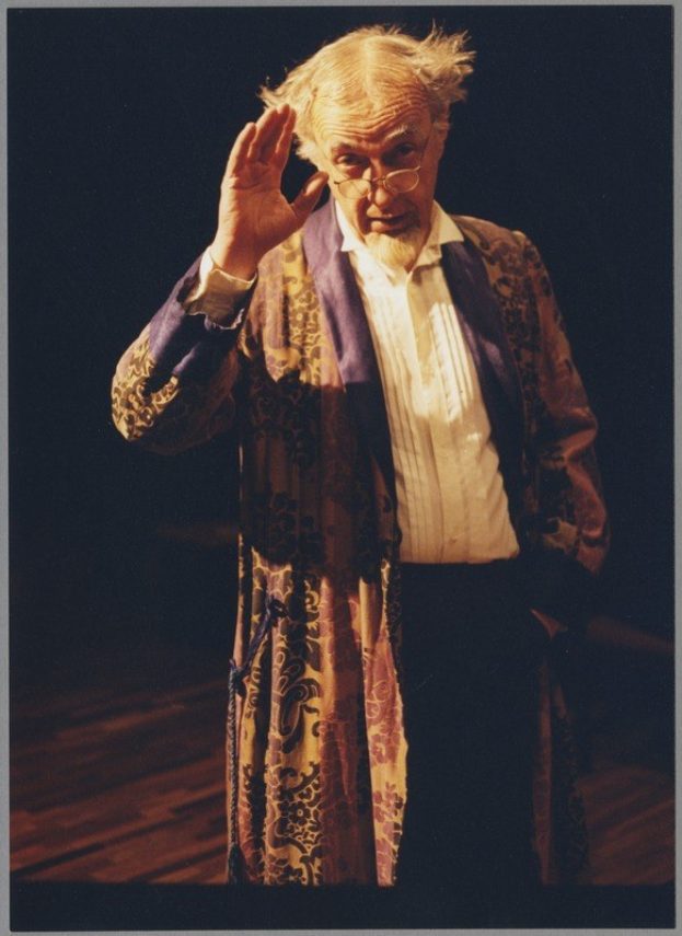 Ein Mann in dunkler Robe und Brille steht auf einer Bühne mit gefalteten Händen vor einem Publikum gegen einen dunklen Hintergrund.