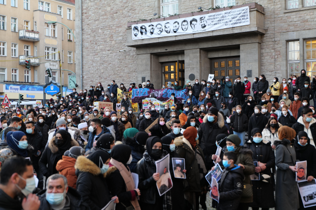 Eine große Gruppe von Menschen in Masken und mit Schildern versammelt sich vor einem Gebäude mit Fenstern, Türen, Bannern, Laternenmasten und Verkehrszeichen, um gegen eine Regierungsentscheidung zu protestieren, Masken zu verbieten.