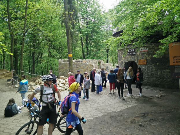 Menschen auf einem Waldweg, einige tragen Helme und Taschen oder Fahrräder, mit Bäumen und einer Steinmauer im Hintergrund und einem Schild, das den Eingang zum Szyb Stylweste Mountainbike-Weg anzeigt.