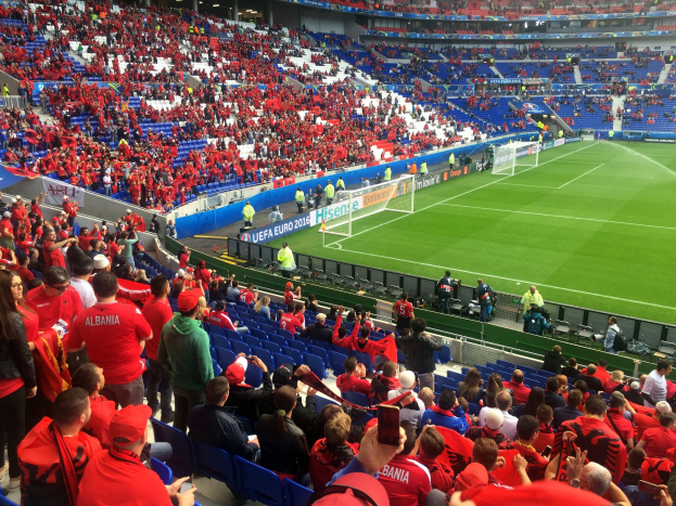 Ein volles Stadion mit sitzenden und stehenden Zuschauern, Bannern, einem Tor und Deckenleuchten, während die Menge gespannt auf ein Fußballspiel wartet.