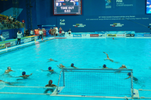 Eine Gruppe von Menschen, die Wasserball in einem Pool mit einem Netz spielen, umgeben von Zuschauern, Tafeln mit Text und Logos, Trägern und einem Bildschirm mit Zahlen und Text bei den 2015 FINA World Championships in Kazan.