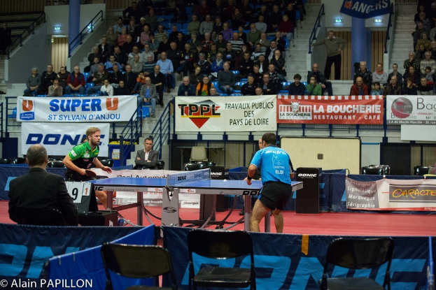 Indoor-Tischtennis-Match mit zwei Spielern, die gegeneinander antreten, Zuschauern, die sitzen und stehen, leeren Stühlen und Werbetafeln in einer Stadionatmosphäre.