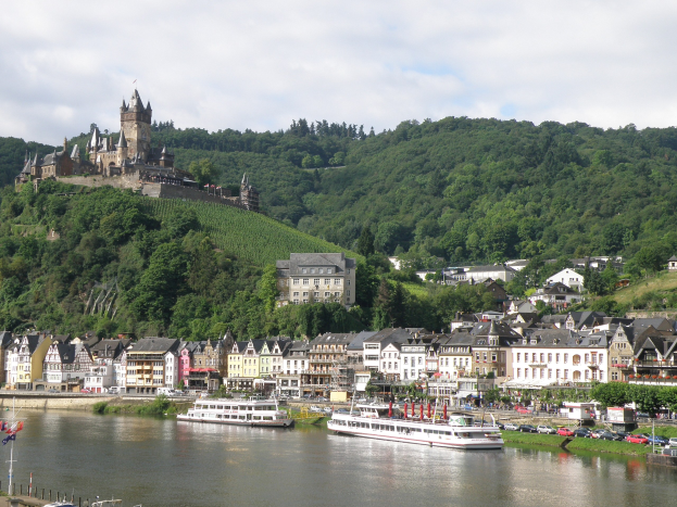 Ein malerischer Blick auf den Rhein in Deutschland mit einer Burg auf einem Hügel im Hintergrund, Booten auf dem Fluss, Fahrzeugen auf einer näheren Straße und einem bewölktem Himmel.