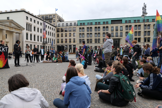 Eine Gruppe von Menschen, die auf dem Boden sitzen und vor einer Menge mit Fahnen und Transparenten eine Rede halten, mit einer Statue und Gebäuden im Hintergrund bei einer Homo-Gegner-Demo in Berlin, Deutschland.
