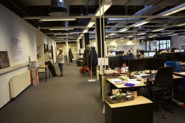 Großes Großraumbüro mit Menschen an Schreibtischen, Computern und Monitoren, umgeben von Papieren, Büchern, Kartons und Wandtafeln mit Text und Bildern, unter Deckenleuchten und in der Nähe von Glasfenstern.