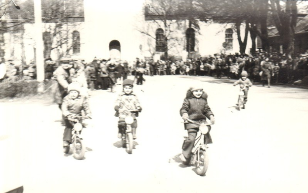 Schwarze-Weiß-Foto von Kindern, die auf Fahrrädern eine Straße hinunterfahren, mit einem Gebäude und Bäumen im Hintergrund.