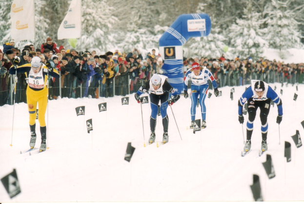 Gruppe von Skifahrern in Helmen, Handschuhen und Skibrillen, die eine schneebedeckte Piste mit Zuschauern und Fahnen im Hintergrund hinunterfahren, einschließlich Text unten.