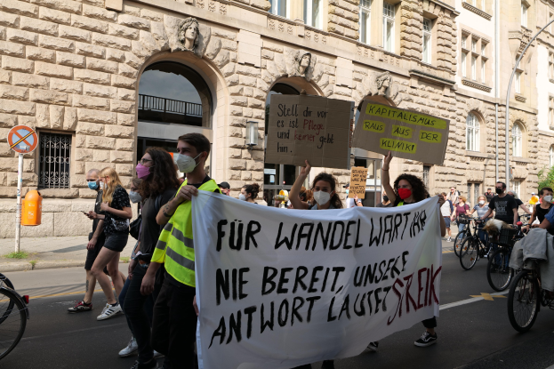 Eine Gruppe von Menschen, die eine Straße entlanggehen, einige halten Schilder und Banner, während andere Fahrräder fahren, mit einem Gebäude im Hintergrund, das Fenster, Bögen, Säulen und Skulpturen aufweist, sowie Bäume und einen Laternenpfahl enthält.