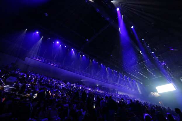 Eine große Menschenmenge sitzt vor einer Bühne mit Deckenbeleuchtung, und auf der rechten Seite ist ein Bildschirm zu sehen.