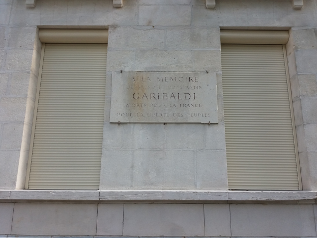 Ein Gebäude mit zwei Fenstern und Läden, das eine Tafel trägt, wahrscheinlich ein Denkmal.