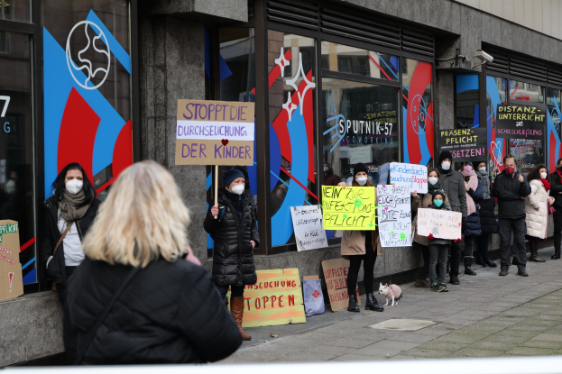 Maskierte Menschen mit Schildern protestieren vor einem Gebäude mit Glaswänden und -türen, auf denen Schilder mit Text und Logos zu sehen sind, während Kameras und ein Hund im Bild sind.