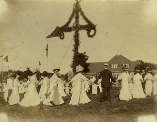 Schwarz-weiß-Foto von Menschen, die um einen Weihnachtsbaum tanzen, mit einem Mann und einer Frau im Vordergrund und Bäumen, Fahnen, einem Gebäude und Himmel im Hintergrund.