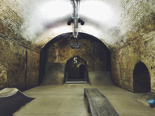 Ein Skatepark mit einem Backstein-Tunnel, einer Rampe, einer Bank und einem beleuchteten Bereich mit einem Schild.