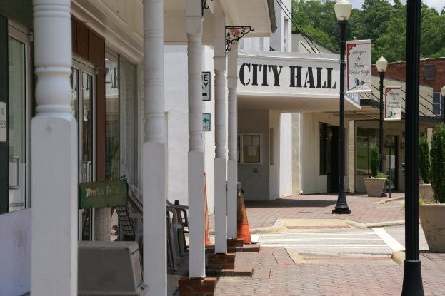 Rathausgebäude mit umgebender Infrastruktur, darunter Straßenmasten, Laternen, Namensschilder, Fahrzeuge, Verkehrskegel, Stühle, Topfpflanzen, Bäume und eine bewölkte Himmel.