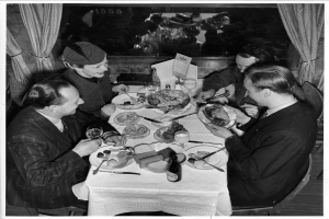 Eine Gruppe von Menschen sitzt an einem Tisch mit Tellern, Gläsern, Flaschen und Besteck, mit Vorhängen im Hintergrund.