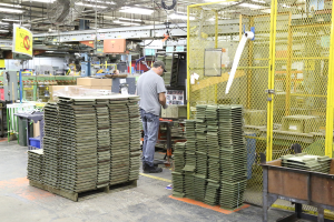 Ein Mann in einer Kappe und einem T-Shirt steht in einer Fabrik und arbeitet in der Nähe von Maschinen auf dem Boden und an der Decke, wobei auch Lüfter und Lampen zu sehen sind.