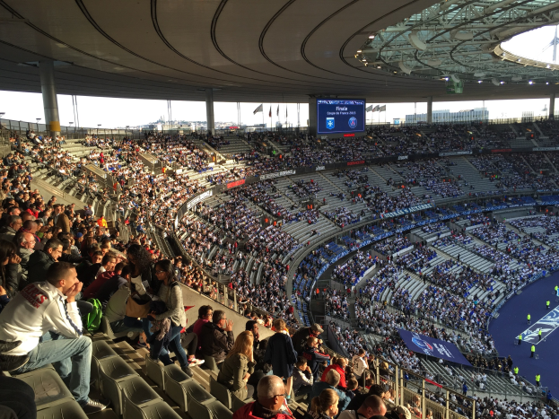 Große Zuschauermenge im Allianz Arena Stadion bei einem Fussballspiel, mit einer Bühne auf der rechten Seite, Fahnen, Stangen und einem Bildschirm im Hintergrund unter einem sichtbaren Himmel.
