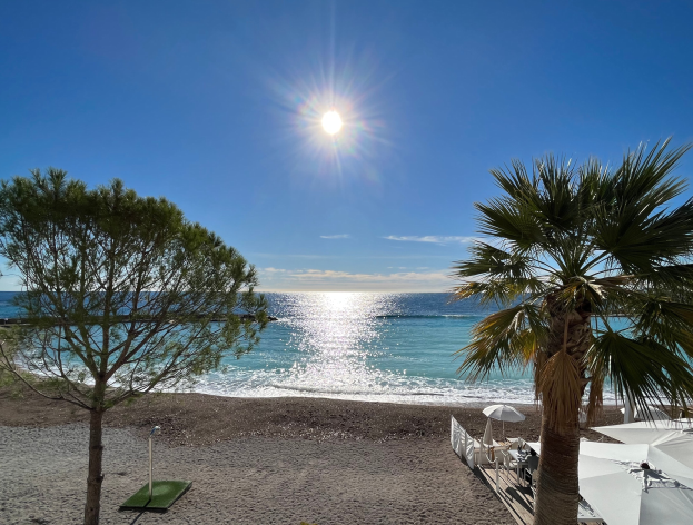 Ein sonniger Strand mit Palmen, Schirmen und üppiger Vegetation, vor einem strahlend blauen Himmel an der französischen Riviera.