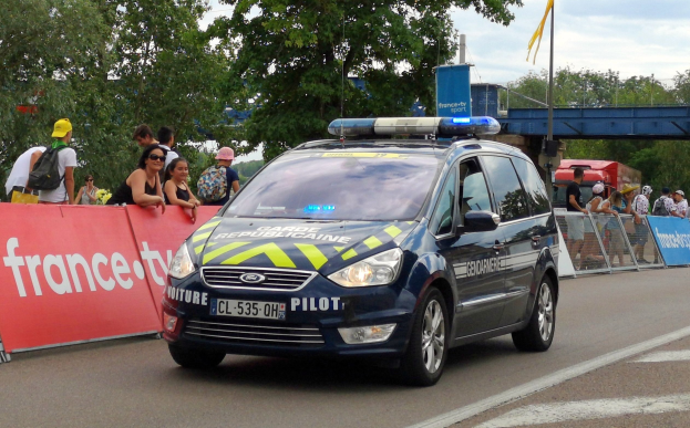 Polizeiauto fährt auf einer Straße neben einer Menschenmenge, mit einem Banner auf der linken Seite, Geländern mit Bannern dahinter und Bäumen, einer Brücke, einer Fahne und Wolken im Hintergrund.