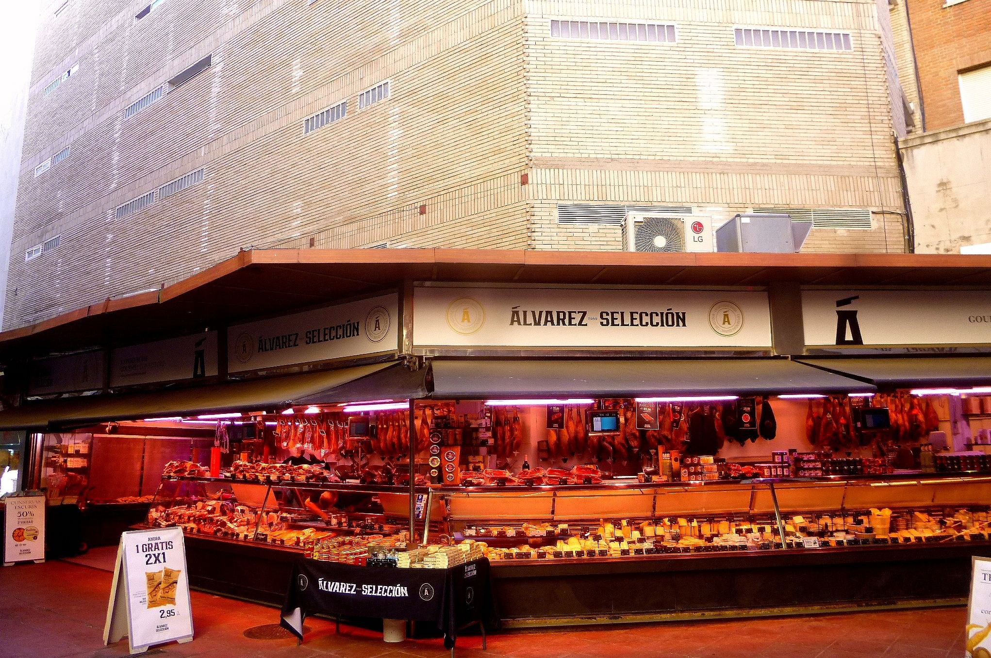 Ein belebter Markt mit verschiedenen Lebensmitteln, die unter Lichtern präsentiert werden, umgeben von Texttafeln, mit Gebäuden und einem klaren blauen Himmel im Hintergrund.