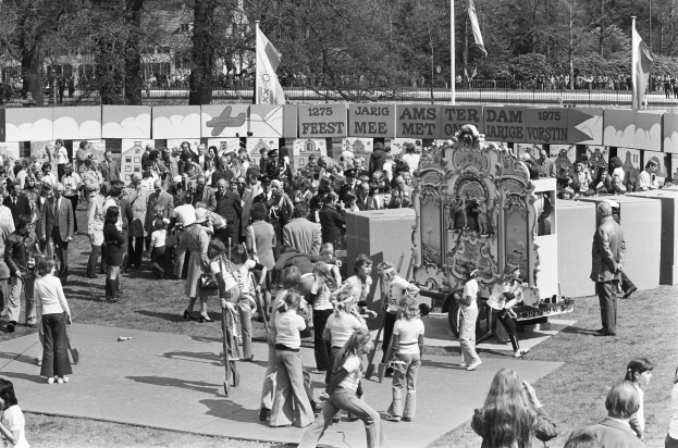 Ein Schwarz-Weiß-Bild einer Menge, die sich um einen mit Fahnen und Bannern geschmückten Karnevalswagen versammelt hat, der in einem Park mit Bäumen und Gebäuden im Hintergrund steht.