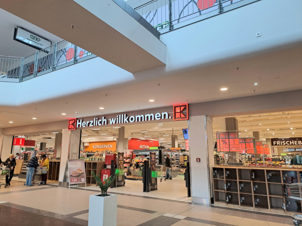 Innenansicht eines Einkaufszentrums mit Menschen, Geschäften, Informationsschildern mit der Aufschrift 'Herzlich willkommen', Beleuchtung, Geländern und einem Topf mit einer Pflanze am Boden.