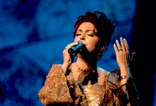 Frau in goldnem Kleid, singend ins Mikrofon auf der Bühne beim Eurovision Song Contest, mit blauem und schwarzem Hintergrund.
