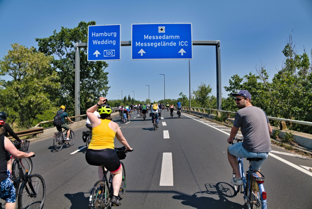 Radfahrer mit Helmen fahren auf einer Straße mit einer Begrenzung und Bäumen auf beiden Seiten unter einem klaren blauen Himmel mit Laternen im Hintergrund, wie durch eine Tafel oben auf dem Bild angezeigt.