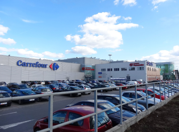 Großes Autohaus mit zahlreichen geparkten Autos, Straßenlaternen, Schildern und Namensschildern, umgeben von Gebäuden und Fahrzeugen auf der Straße unter einem bewölkten Himmel.