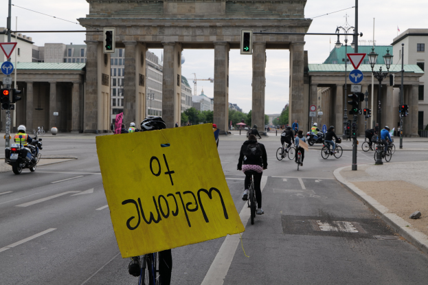 Eine Gruppe von Radfahrern in Helmen fährt vor dem Brandenburger Tor in Berlin, Deutschland, vorbei, während einer unter einem klaren blauen Himmel ein gelbes Schild hält.