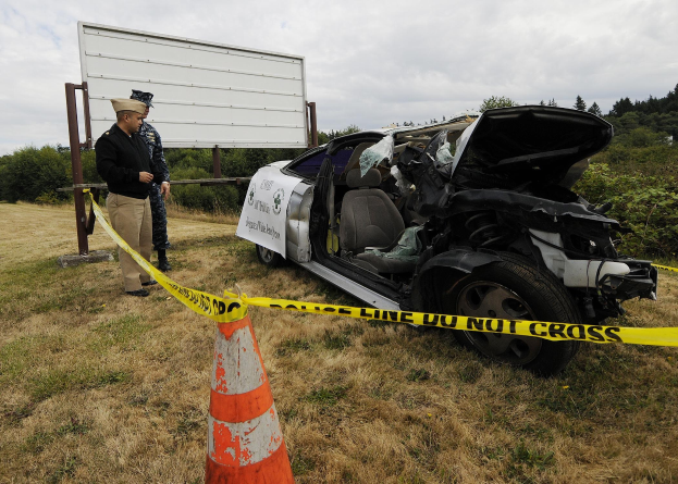 Ein Polizist in Uniform steht neben einem schwer beschädigten Auto an der Straße, mit einem Verkehrskegel und einem Band an einem Pfahl in der Nähe und Bäumen und einem bewölkten Himmel im Hintergrund.