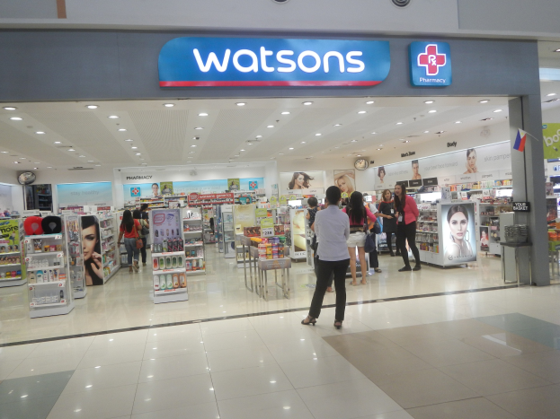 Eine Watsons Apotheke in einem Einkaufszentrum mit Menschen, Regalen, Schildern, einer Wanduhr und Deckenbeleuchtung.