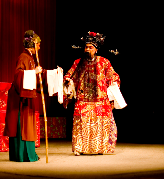 Ein Mann in rotem Gewand hält einen Stock und Papiere auf der Bühne und steht neben einer Frau in grünem Kleid, mit einem roten Vorhang dahinter.